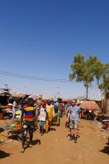 A diverse group of people are walking down a dirt road lined with market stalls under a clear blue sky in Niamey, Niger.