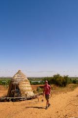 A man walks on a dirt path in Niger, near a traditional straw hut, under a clear blue sky.