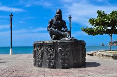 Parque Nacional Natural Tayrona Tour de 2 días desde Cartagena A statue of a seated man on a carved stone base stands on a brick plaza overlooking the ocean under a bright blue sky.
