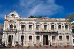 Parque Nacional Natural Tayrona Tour de 2 días desde Cartagena An old building with a dome, balconies, and ornate decorations is set against a blue sky with clouds. People walk on the sidewalk in front.