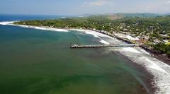 Aerial shot of El Sunzal beach in El Salvador, featuring a pier stretching into the ocean, a shoreline of black sand, and lush green vegetation lining the coast.