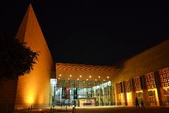 Illuminated by warm lights, a modern building with a pyramid-like structure stands against a dark night sky, with two people walking in front.