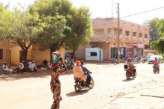 A vibrant street scene in a sunny African town, with locals socializing, motorbikes passing by, and a woman carrying a load on her head.