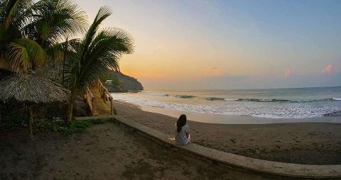 A serene beach scene at sunset features a woman sitting on a low wall, gazing at the ocean waves, framed by palm trees and a thatched umbrella.