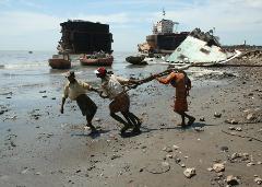 Three men pull a thick cable on a beach littered with debris from shipbreaking, with hulks of ships in the background.