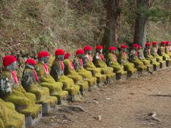 A row of moss-covered stone statues wearing red hats and bibs sit in a line along a path in a forest.