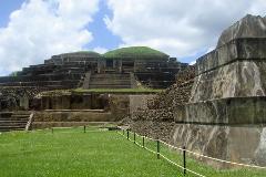 A layered stone pyramid with a grassy top stands amidst green grass, partially blocked by a rope barrier.