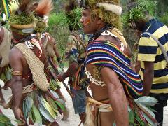 A group of people, some in traditional clothing with leaf skirts and headwear, are walking outdoors. One man in the foreground has a colorful shawl and shell necklace.