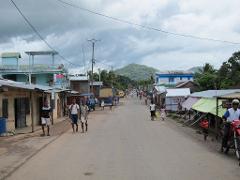 This is a street view of a town in Madagascar, with people walking along the main road flanked by small shops and buildings under a cloudy sky.