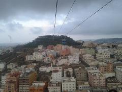 A high-angle shot of a city with dense buildings, a mountain with green lights, and a gondola cable car running through a cloudy sky.