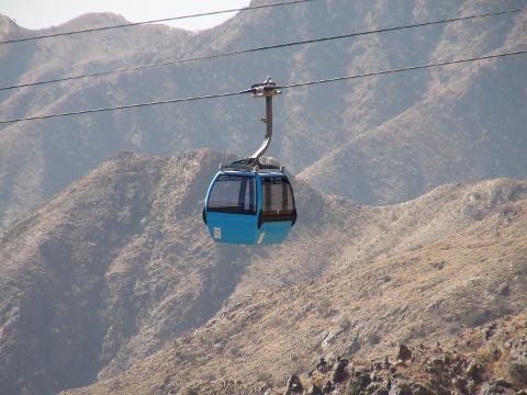 A blue cable car hangs suspended above a rugged, mountainous landscape under a hazy sky.