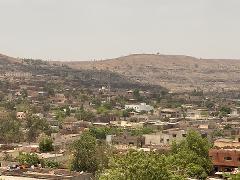 A high-angle shot reveals a densely populated town nestled beneath a brown, arid hill, with a hazy sky overhead.