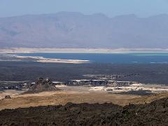 A scenic landscape featuring a stark contrast between dark, volcanic terrain and a vibrant blue lake under hazy mountains.