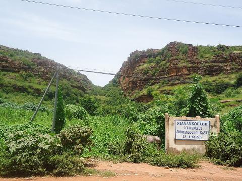 A lush green valley with rocky cliffs on either side frames a sign that reads "NIANANKOULOU ICI DISPARUT SOUMANGOUROU KANTE EN 1235" surrounded by dense vegetation, with a utility pole and wires in the background.