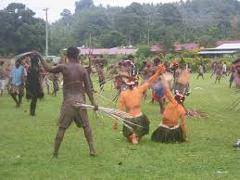 Certainly! Here is a description of the image in one sentence and under 320 characters:The image shows a group of people in Papua New Guinea, possibly performing a traditional dance or ritual, on a grassy field against a backdrop of buildings and foliage.