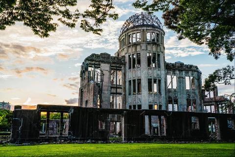 The Hiroshima Peace Memorial, also known as the Atomic Bomb Dome, stands amidst green grass and framed by trees under a sky with pastel clouds, a solemn reminder of the past.