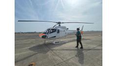 A white helicopter with an orange nose sits on a tarmac with a person standing nearby under a partly cloudy sky.
