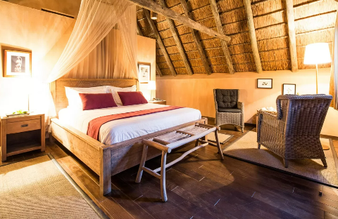 Cozy bedroom with a wooden bed featuring a canopy, an end-of-bed bench, and two chairs on a rug under a thatched roof.