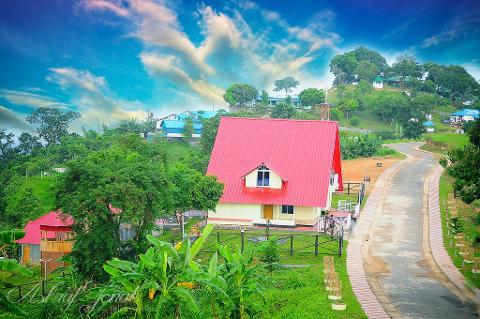 A scenic view showcases a house with a red roof amid lush greenery and a winding road on a hillside, under a bright sky with scattered clouds.