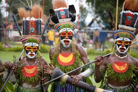 Three men in traditional Papua New Guinea attire with vibrant face paint, feather headdresses, and grass skirts stand holding spears and a long wooden instrument.