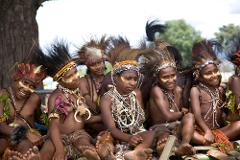 A group of young Papua New Guinean people in traditional attire, including face paint, headdresses, and necklaces, sit together.