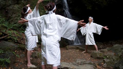 Three people wearing white robes and headbands are standing in front of a waterfall, with their arms outstretched.