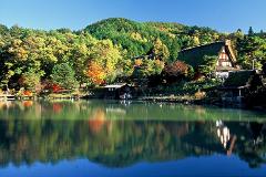 A picturesque Japanese landscape features a tranquil lake reflecting autumn trees and traditional thatched-roof houses nestled against a green hillside under a clear blue sky.