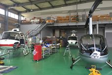 Aquí hay una descripción de la imagen: Una toma interior de un almacén/garaje que contiene tres pequeños helicópteros en reparación o mantenimiento, con herramientas y equipos esparcidos sobre un piso verde.