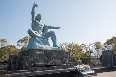A statue of a man sitting cross-legged with one arm raised in the air stands on a stone platform surrounded by trees, with flowers in the foreground.