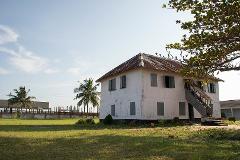 An old, two-story white building with a rusty roof stands on a grassy lawn, with palm trees and another building visible in the background, under a partly cloudy sky.