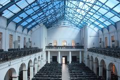 A large, bright auditorium with rows of black chairs under a glass ceiling and a balcony supported by arches and columns.