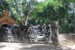 An artistic gate with elaborate carvings depicting figures and symbols, standing amidst lush green trees in a natural setting.