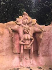 A pinkish-hued outdoor sculpture depicts a nude figure with a cap and arms crossed, flanked by a draped figure with arms outstretched under an abstract headpiece.
