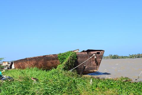 Salamanca Island Road Park Birdwatching Tour from Santa Marta A rusty abandoned boat sits on a grassy riverbank under a clear blue sky, with lush greenery and a distant shoreline visible in the background.