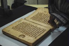 An old book with Arabic writing lies open on a table, with a person's hand partially visible in the right side of the frame.