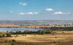 A scenic view of a lake with dead trees rising from the water, a grassy plain in the foreground, and a town nestled in the distance under a blue sky with scattered clouds.