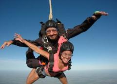 A tandem skydiving pair, strapped together and wearing helmets and goggles, smiles broadly as they freefall against a clear blue sky.