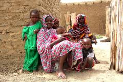 A group of African women and children are gathered together in front of a mud brick building.