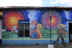 A colorful building mural in El Salvador features stylized cats, mandala-like designs, and the text "Hecho en El Salvador Ataco," advertising textiles and arts.