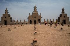 The image depicts a view of the Great Mosque of Djenné in Mali, with people in colorful attire walking on the sandy surface, surrounded by conical structures.