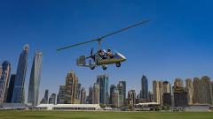 A silver gyrocopter flies in front of a city skyline on a clear day, with two people in the cockpit and a green field below.