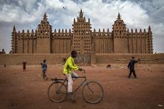 A boy rides a bicycle in front of the Great Mosque of Djenné in Mali, surrounded by other people and a dusty landscape.