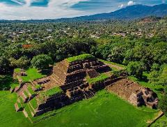 Aerial view of the pre-Columbian Maya archaeological site of Tazumal, with its stepped pyramid structures amidst green lawns, surrounded by lush trees and a distant mountain backdrop.