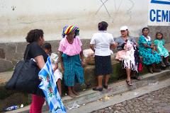 A group of women and children are gathered on a curb near a wall, with one woman holding a blue patterned cloth bag and another wearing a colorful head wrap.