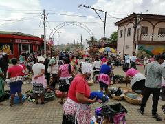 A bustling open-air market scene with vendors and shoppers, many wearing masks, selling and buying produce and goods.