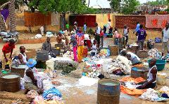 A group of people are washing clothes outdoors, using barrels and a communal washing area, surrounded by piles of colorful fabrics.