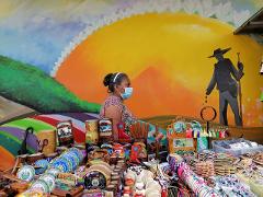 A woman wearing a mask stands behind a vibrant vendor table filled with souvenirs against a colorful mural backdrop.