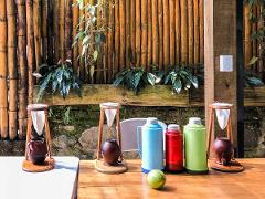 Three wooden coffee makers, colorful thermoses, and a lime sit on a wooden table against a background of bamboo and greenery.