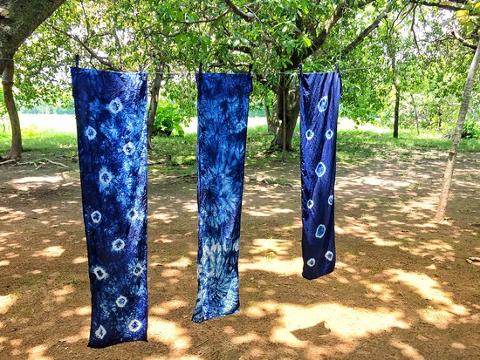 Three blue tie-dye fabrics hang on a clothesline under trees, casting shadows on the ground.