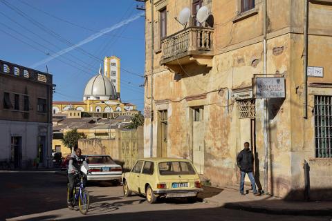 A street view in Asmara, Eritrea with yellow buildings, two cars, a cyclist, a man walking out of a building, and a church in the background.